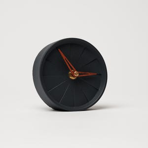 Peut inclure: Une petite horloge ronde noire au design unique. Le cadran de l'horloge présente un motif radial et des aiguilles orange et un centre doré. L'horloge est un objet décoratif.