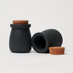 Può includere: Due vasetti in ceramica nera con tappi di sughero. I vasetti sono rotondi e hanno una forma leggermente bombata. I tappi sono in sughero naturale.