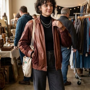 May include: A brown leather jacket with dark brown textured knit sleeves, worn over a black turtleneck sweater and dark gray corduroy pants. A cream-colored canvas tote bag hangs from the shoulder, and a silver chain necklace is worn.