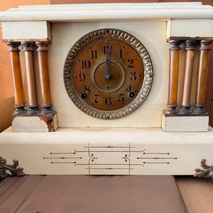 May include: A cream-colored mantel clock with a central circular face featuring black Arabic numerals and dark hands. The clock is flanked by two sets of three amber-toned columns and features decorative black line detailing on the base.