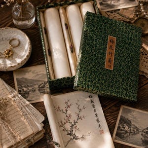 May include: A rectangular green box with a patterned texture and a label featuring Chinese characters, containing several rolled scrolls. One scroll is unrolled, displaying a painting of a flowering plum branch and vertical Chinese calligraphy.