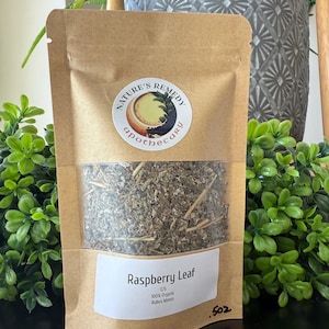 May include: A brown paper bag with a clear window, filled with dried raspberry leaves. The bag has a white label with the text "Raspberry Leaf" and a circular sticker with the words "Nature's Remedy Apothecary". The bag contains .5 oz of product.