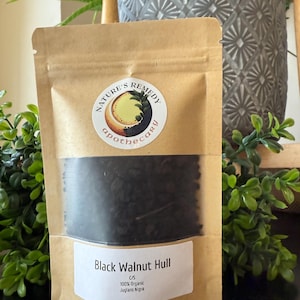 May include: A brown paper bag with a clear window, filled with black walnut hull. The bag has a label with the text "Black Walnut Hull", "100% Organic", and "Juglans Nigra". The bag also has a circular sticker with the text "Nature's Remedy apothecary".