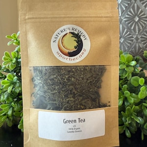 May include: A brown paper bag with a clear window, filled with green tea leaves. The bag has a white label that reads "Green Tea" and "100% Organic Camellia Sinensis." A circular sticker with the words "Nature's Remedy Apothecary" is also visible.
