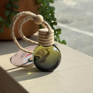 May include: A small, round, olive-green glass air freshener bottle with a wooden cap and a natural rope loop. The bottle has a label that reads "ISCES". The rope is adorned with gold flecks. The bottle is sitting on a light-colored surface.