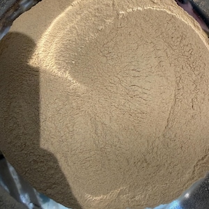 Puede incluir: Primer plano de un polvo marr&oacute;n claro que llena un cuenco de acero inoxidable. El polvo tiene un aspecto fino y texturizado. La superficie brillante y met&aacute;lica del cuenco contrasta con el polvo mate.