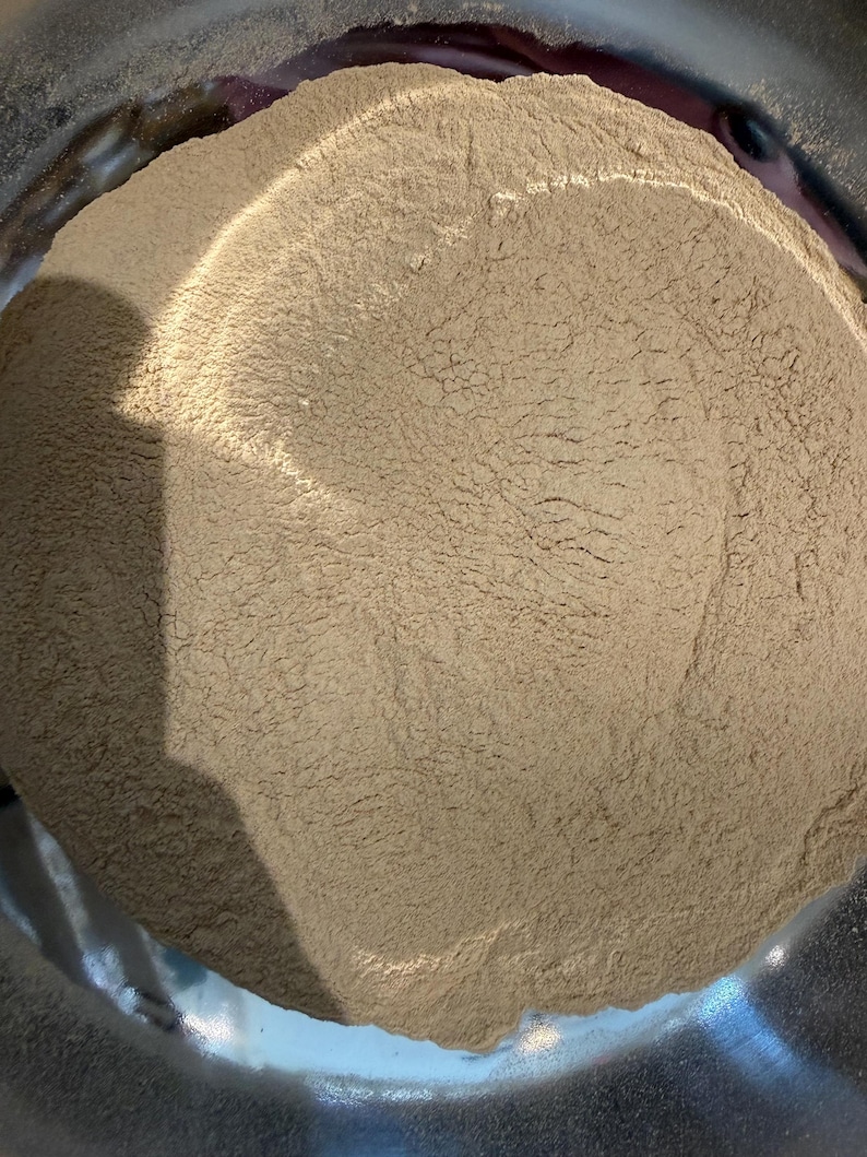 Puede incluir: Primer plano de un polvo marr&oacute;n claro que llena un cuenco de acero inoxidable. El polvo tiene un aspecto fino y texturizado. La superficie brillante y met&aacute;lica del cuenco contrasta con el polvo mate.