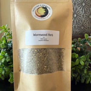 May include: A brown paper bag with a clear window, filled with dried wormwood herb. The bag has a white label with the text "Wormwood Herb" and "100% Organic Artemisia Absinthium." The bag also has a circular sticker with the text "Nature's Remedy Apothecary."