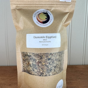 May include: A brown paper bag with a clear window showing dried chamomile flowers. The bag is labeled "Chamomile (Egyptian)" and "Nature's Remedy Apothecary". The bag contains 1.5 ounces of chamomile and costs $15.99.