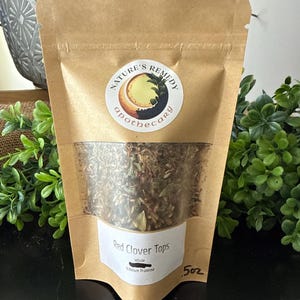 May include: A brown paper bag with a clear window, filled with dried red clover tops. The bag has a white label with the text "Red Clover Tops" and "5oz". A circular sticker with the words "Nature's Remedy Apothecary" is on the bag.