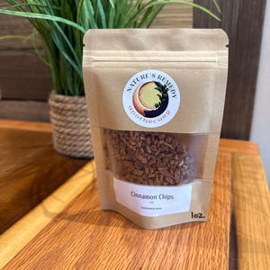 May include: A brown kraft paper pouch containing small, dried cinnamon chips. The label features the text Nature's Remedy Apothecary and Cinnamon Chips, with a handwritten 1oz marking. The pouch has a clear window revealing the brown spice pieces.