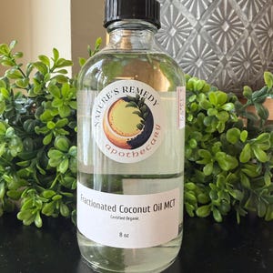 Op de afbeelding: Een heldere glazen fles Fractionated Coconut Oil MCT van Nature's Remedy Apothecary. De fles heeft een zwarte dop en een wit etiket met tekst en een logo. De fles bevat 237 ml gecertificeerde biologische olie.