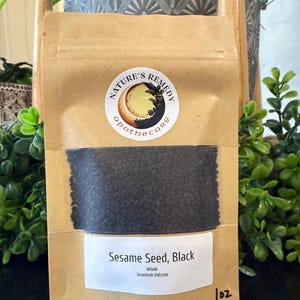 May include: A brown paper bag filled with black sesame seeds. The bag has a white label with the text "Sesame Seed, Black" and "Whole Sesamum Indicum". A round sticker with the words "Nature's Remedy apothecary" is on the bag. The bag is labeled "1 oz".