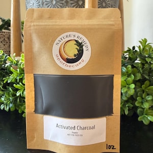 Może przedstawiać: Brązowa papierowa torebka z proszkiem węgla aktywnego. Torebka ma przezroczyste okienko ukazujące czarny proszek. Torebka ma białą etykietę z napisem "Activated Charcoal" i "NOT FOR FOOD USE". Torebka jest oznaczona jako "1oz" i ma okrągłą naklejkę z napisem "Nature's Remedy Apothecary".
