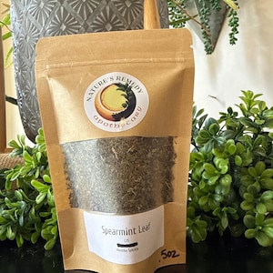 May include: A brown paper bag with a clear window, filled with dried spearmint leaves. The bag has a white label that reads "Spearmint Leaf" and "5oz". A circular sticker with the words "Nature's Remedy apothecary" is also on the bag.