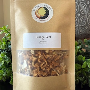 May include: A brown paper bag with a clear window containing dried orange peel. The bag has a label that reads "Orange Peel" and "100% Organic Citrus x sinensis". The bag also has a circular sticker with the words "Nature's Remedy apothecary".