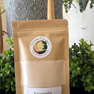 Puede incluir: Una bolsa de papel marr&oacute;n con una ventana transparente que contiene un polvo beige. La bolsa tiene una etiqueta blanca que dice "Ashwagandha Powder" y una pegatina circular con las palabras "Nature's Remedy Apothecary". El n&uacute;mero "202" est&aacute; impreso en la parte inferior derecha.