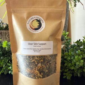 May include: A brown paper bag of herbal tea blend labeled "Clear Skin Support" with the apothecary logo. The bag contains a mix of dried herbs and flowers. The text on the bag lists ingredients like burdock root, dandelion root, and chamomile. The bag is 2 oz.