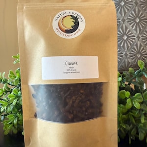 May include: A brown paper bag with a clear window, filled with whole cloves. The bag has a white label that reads "Cloves" and "100% Organic". The bag also has a circular sticker with the words "Nature's Remedy Apothecary". The bag is labeled "2 oz".