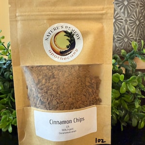Könnte beinhalten: Ein brauner Papiertüte mit einem transparenten Fenster, das Zimtstücke enthält. Der Beutel hat ein weißes Etikett mit der Aufschrift "Cinnamon Chips" und "100% Organic Cinnamomum verum". Ein Aufkleber mit "Nature's Remedy apothecary" ist ebenfalls sichtbar.
