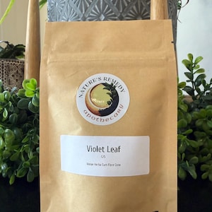 May include: A brown paper bag with a white label that reads "Violet Leaf" and "Nature's Remedy apothecary" in a circular logo. The bag also has the text ".5 oz" in the bottom right corner. The bag is surrounded by greenery.