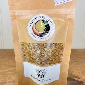 May include: A brown paper stand-up pouch containing yellow and orange bee pollen granules. The label features the text Nature's Remedy apothecary and Bee Pollen Granules with a bee illustration. A white price tag with $8.99 is attached to the top.