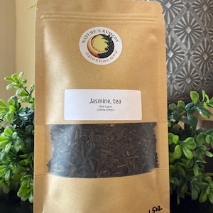 Op de afbeelding: Een bruine papieren zak met losse jasmijnthee. Een transparant venster toont de theebladeren. Het etiket luidt "Jasmine, tea" en "100% Organic". De zak heeft een ronde sticker met de tekst "Nature's Remedy apothecary".