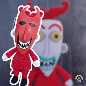 Pode incluir: Boneca vermelha de crochê de um personagem com máscara diabólica, chifres e um sorriso largo e dentuço. A boneca está vestindo uma roupa vermelha com sapatos pretos e luvas brancas. O fundo apresenta uma imagem borrada de um personagem semelhante.