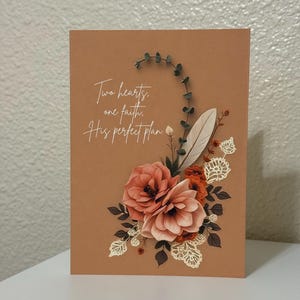 May include: A greeting card with a brown background features floral arrangements in shades of peach and orange, with a white feather and lace accents. The card has the text "Two hearts, one faith, His perfect plan."