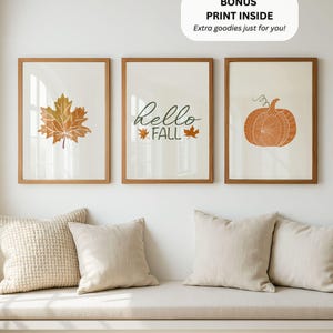 Może przedstawiać: Trzy oprawione w drewniane ramy plakaty ścienne. Lewy plakat przedstawia pomarańczowy liść klonu, środkowy zawiera napis "Hello FALL" z dwoma małymi listkami, a prawy prezentuje ilustrację pomarańczowej dyni.
