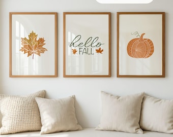 May include: Three framed wall art prints in wooden frames. The left print shows an orange maple leaf, the center print features the text "Hello FALL" with two small leaves, and the right print displays an orange pumpkin illustration.