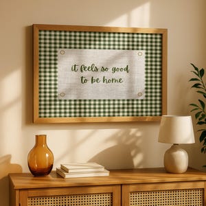 May include: A framed wall art piece with a green and white checkered pattern and the text "it feels so good to be home". The frame is made of wood. A brown glass vase, books, and a lamp sit on a wooden cabinet.