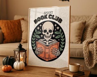May include: A framed print with a gold border showing a skull reading a book, encircled by a black design with the words "Spooky Book Club." The image is on a wooden table with pumpkins, a lantern, and a coffee cup.