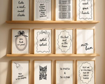 May include: A set of 12 wall art prints displayed on three wooden shelves. Each print features black text and graphics on a white or pinstriped background, including phrases like take a seat, guest check, shower karaoke, wash your hands, and splish splash.