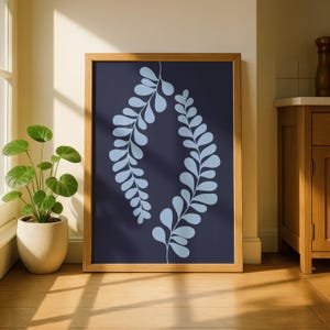 May include: A framed art print featuring a navy blue background with a light blue leaf design. The artwork is encased in a natural wood frame and leans against a wall. The print is displayed on a wooden floor, with a potted plant visible.