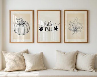 May include: Three framed wall art prints with light beige backgrounds. The left print features a black line drawing of a pumpkin. The center print displays the text "hello FALL" with two leaf icons. The right print shows a black line drawing of a maple leaf.