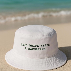 May include: White bucket hat with the text "THIS BRIDE NEEDS A MARGARITA" in green. The hat is sitting on a sandy beach with the ocean in the background. A fun accessory for a bachelorette party or bridal event.