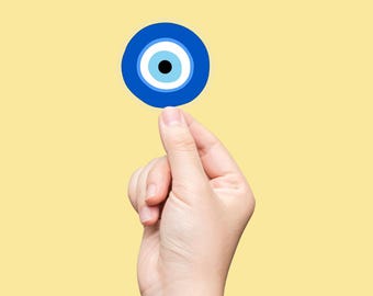 May include: A blue and white evil eye charm with a black center, held between fingers against a yellow background. The charm is round and features concentric circles of blue, white, and black.