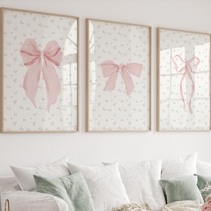 Puede incluir: Tres impresiones artísticas enmarcadas con lazos rosas de acuarela sobre un fondo floral claro. Las impresiones se exhiben sobre un sofá blanco con cojines decorativos. Los marcos son de color madera clara.