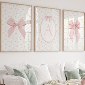 Puede incluir: Tres impresiones enmarcadas con lazos rosas y la letra "A" sobre un fondo floral. Las impresiones están en marcos de madera clara, creando una estética suave y femenina. La obra de arte se exhibe sobre un sofá blanco con cojines decorativos.