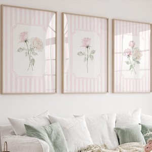 Può includere: Tre stampe artistiche floreali incorniciate. Ogni stampa presenta un'illustrazione di rosa ad acquerello in una cornice decorativa su uno sfondo a righe rosa e bianche. Le cornici sono di colore legno chiaro. Le stampe sono esposte sopra un divano bianco.
