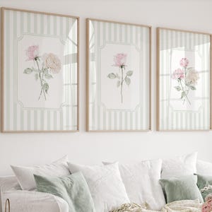 Puede incluir: Tres impresiones artísticas florales enmarcadas. Cada impresión presenta una ilustración de rosa en acuarela en tonos rosa y crema. El fondo tiene un patrón de rayas verdes y blancas claras, y los marcos son de color madera clara.