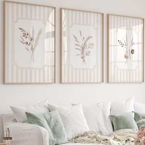 May include: Three framed botanical prints with beige and white striped backgrounds. Each print features a different arrangement of dried flowers in shades of brown, beige, and purple. The frames are a light wood colour, and the prints are displayed above a white sofa with green and white cushions.