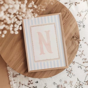 May include: A framed print featuring a large, pink letter "N" on a white background with a scalloped border. The background has light blue and white vertical stripes. The frame is made of light-colored wood.