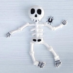 May include: A white felt skeleton with black embroidered details, lying on a white wooden surface. A small black felt spider with white eyes is perched on the skeleton's arm.