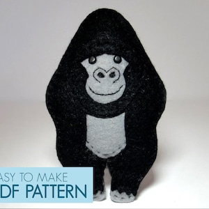 以下が含まれることがあります： 黒と灰色のフェルトで作られたゴリラのぬいぐるみです。ゴリラは腕を体の横にまっすぐに立てています。おもちゃは黒い目で縫い付けられた笑顔の優しい顔をしています。画像の下部に「EASY TO MAKE PDF PATTERN」というテキストがあります。
