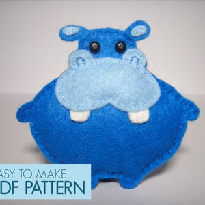 Op de afbeelding: Een handgemaakt blauw vilten nijlpaardspeelgoed met een lichtblauwe snuit en oren en witte tanden. Het nijlpaard heeft zwarte knoopogen en een schattige, ronde vorm. De tekst "EASY TO MAKE PDF PATTERN" staat onderaan.