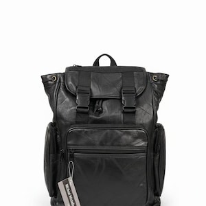 May include: Black leather backpack with multiple compartments, including side pockets and a front zippered pocket. Features adjustable straps, a top handle, and a flap closure with buckles. The tag reads "Michael Michelle".