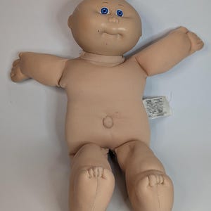 以下が含まれることがあります： ビニール製の頭部、明るい肌色、鮮やかな青い目を持つソフトボディの人形。人形は短い明るい色の毛糸の髪、小さなへそ、広げた腕が特徴。側面にテキストが印刷された白い布製のタグが付いている。