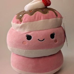 Puede incluir: Un juguete de peluche de postre de tres niveles en tonos rosa, blanco y marrón. La capa superior presenta una crema blanca y una fresa roja. La capa intermedia tiene una cara sonriente con pestañas negras y mejillas rosadas. Una etiqueta cuelga del costado.